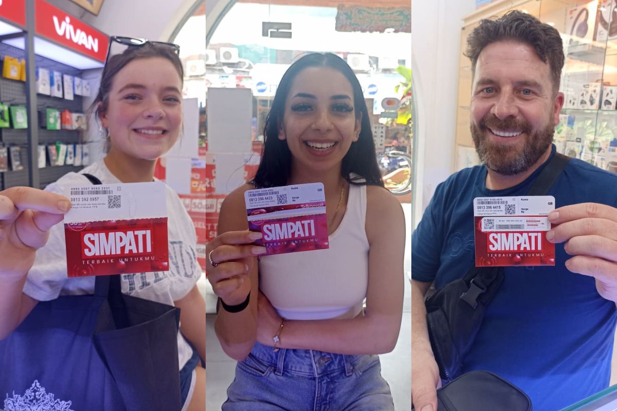 Three people smiling and holding up SIMPATI travel cards inside Balitel's store. The Telkomsel SIMPATI brand name is clearly visible on the red and white cards they are holding.